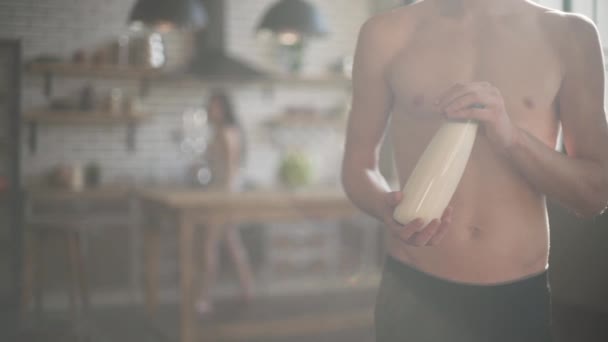 Άνθρωπος με ένα γυμνό κορμό κρατώντας ένα μπουκάλι γάλα στέκεται στην κουζίνα κάτω από τον ήλιο. Χαριτωμένο κορίτσι με τα νυχτικά έρχεται με τον τύπο της, παίρνει το γάλα και φύλλα. — Αρχείο Βίντεο