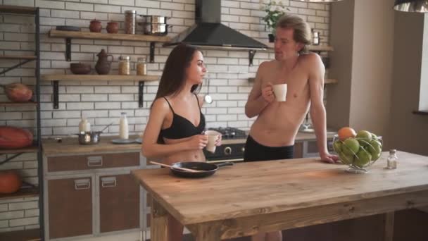 Fresado joven pareja semidesnuda en la hermosa cocina loft. Guy y una chica en ropa interior están bebiendo su café de la mañana mientras están de pie cerca de la mesa de la cocina a la luz del sol . — Vídeos de Stock