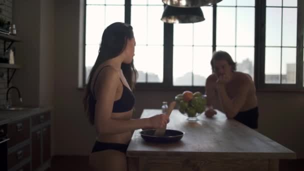 Young lovers cooking breakfast in the kitchen. Brunette in bra and young man in underwear stand near kitchen table. Guy likes his girlfriend and helps her to cook passing the salt. — Stock Video