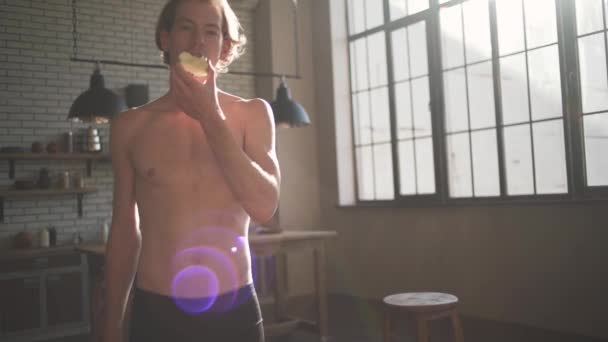 Portret van een knappe man met een ontbloot bovenlijf eten een appel staande in de keuken in het zonlicht. — Stockvideo