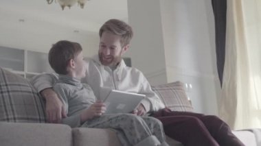 Sakallı baba ve küçük oğlu pencerenin geniş oturma odası ve oyun tablet kanepede oturuyor. Baba çocuğunu aracını kullanabilmek için öğretir. Mutlu baba ve çocuk. Baba-oğul ilişkisi.