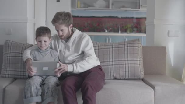 Padre y niño barbudo usando tableta digital sentados en el sofá en la sala de estar grande. Papá enseña a su hijo. Relación padre-hijo . — Vídeos de Stock