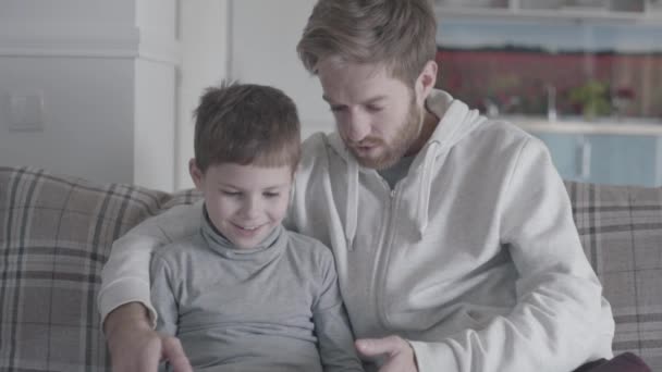 아버지와 큰 거실에 태블릿에 소파에 앉아 작은 아들. 아빠는 그의 아이 가르치고 있습니다. 아버지-아들의 관계. — 비디오