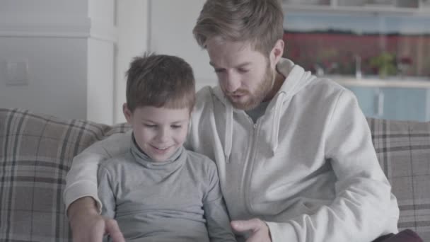 Padre e figlio piccolo seduti sul divano in ampio soggiorno e giocare su tablet. Papà insegna a suo figlio. Relazione padre-figlio . — Video Stock