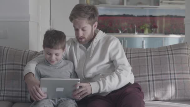 Бородатий батько та дитина, використовуючи цифровий планшет, сидячи на канапі у вітальні, великий. Тато вчить його сина. Щаслива родина провести час разом. Батько син відносин. — стокове відео