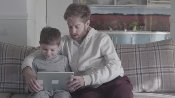 Vousatý otce a dítě pomocí digitálních tabletu sedí na gauči ve velkém obývacím pokoji. Táta učí svého syna. Šťastná rodina trávit čas společně. Vztah otec syn. — Stock video