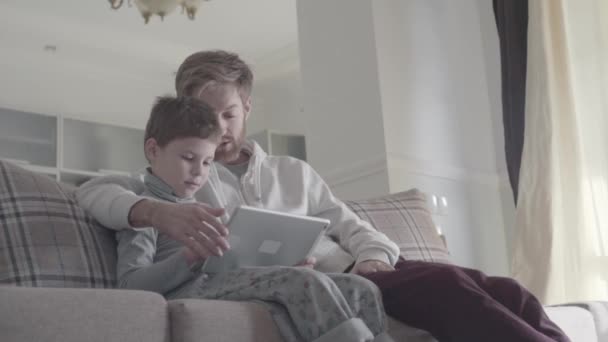 Ojciec i dziecko za pomocą cyfrowego tabletu siedzi na kanapie w duży pokój dzienny. Tatuś uczy swoje dziecko. Szczęśliwy tata i dziecko. Relacji ojciec syn. — Wideo stockowe