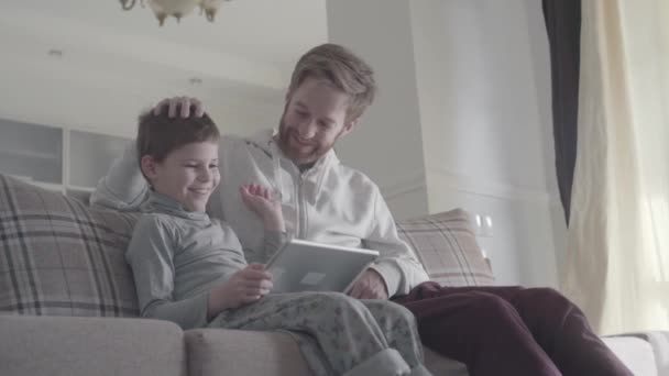 Sonriendo feliz padre e hijo utilizando tableta digital sentado en el sofá en la sala de estar grande. La familia feliz pasa tiempo juntos. Padre acaricia al niño en la cabeza. Relación padre-hijo . — Vídeos de Stock