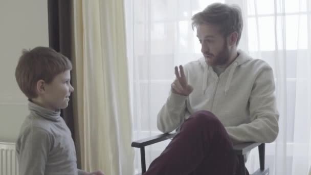 Der bärtige Papa sitzt im Sessel am großen Fenster und spricht emotional mit seinem kleinen Sohn. Der Vater stimmt dem Kind zu. Vater-Sohn-Beziehung. — Stockvideo