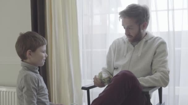 Bearded father sitting in arm-chair near big window and talking with his little son. Father agrees with the child. Father-son relationship. — Stock Video