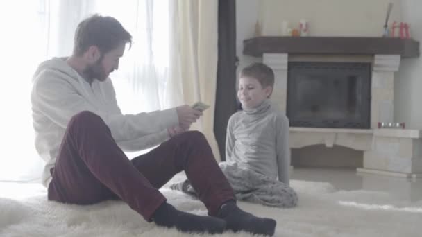 Papá barbudo está sentado con su pequeño hijo en la alfombra. Padre está de acuerdo con el niño. El chico cuenta el dinero. Relación padre-hijo . — Vídeos de Stock