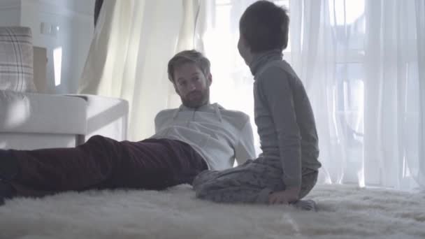 Jonge, bebaarde vader gesprekken met het kind. Bebaarde papa liggen op het witte tapijt en zijn zoon zit naast hem. Vader-kind relatie. — Stockvideo