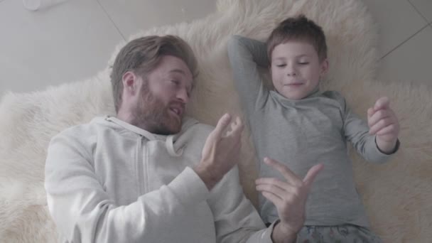 Bebaarde vader en zijn zoontje ligt aan de witte tapijt bont en interessant gesprek hebben. Vader-kind relatie. — Stockvideo