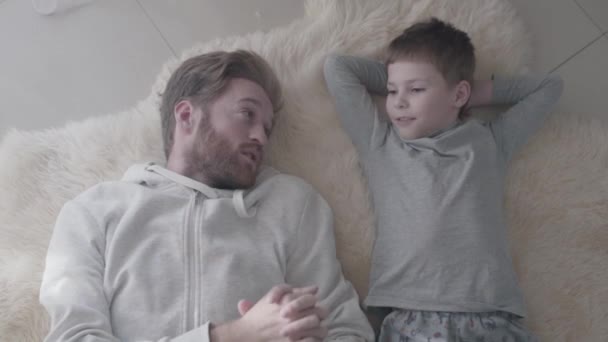 Bearded father and his little son lying at the white fur carpet and have interesting conversation. Father-child relationship. — Stock Video