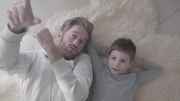 Fun daddy with beard and his little son lying at the white fur carpet and have interesting conversation. Father-child relationship. — Stock Video