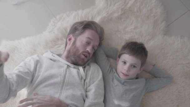 Bearded dad and his little son lying at the white fur carpet and have interesting emotional conversation. Father-child relationship. — Stock Video