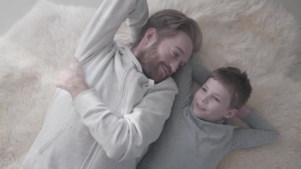 Smilling bearded father and his little son lying at the white fur carpet and have interesting conversation. Father talk with his kid like equal. Father-child relationship. — Stock Video