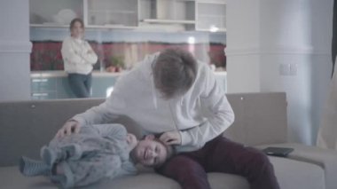 Baba oğlu, gülen çocuk gıdıklıyor. Anne arka planda duruyor. Aile ilişkileri.