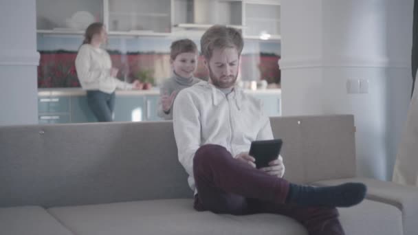 Bebaarde man zittend op de Bank in de woonkamer met tablet. Zoon komt achter zijn vader zittend op de Bank en sluit zijn ogen met zijn palmen. Papa pak hem en begint te spelen met kind, glimlachend. — Stockvideo