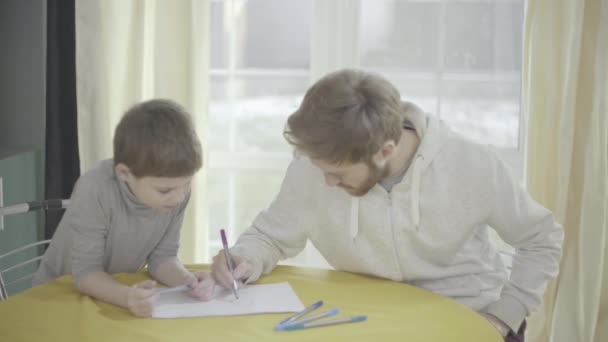 Vousatý otce a jeho malý syn malovat na papír sedí v obývacím pokoji u stolu. Vztah otec dítě. — Stock video