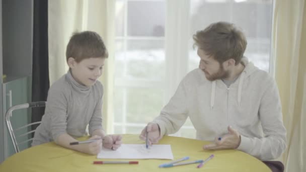 Bebaarde smilling vader en zijn zoontje verf op papier in de woonkamer aan de tafel zit. Vader-kind relatie. — Stockvideo