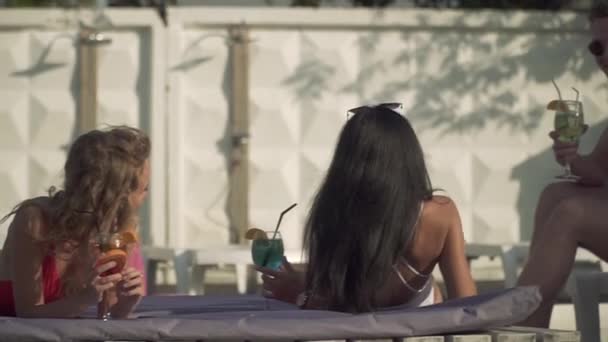 一个穿着泳裤和太阳镜的年轻人和两个穿着泳衣的漂亮女友在室外游泳池附近的日光浴床上交流。男人和两个女孩放松, 喝鸡尾酒. — 图库视频影像