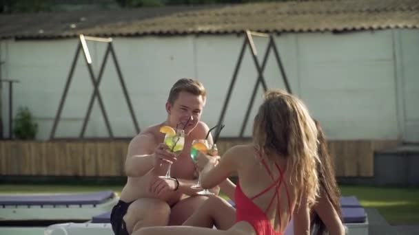 Tres amigos sonríen, hacen clic en vasos con cócteles decorados, y se divierten un día de verano en el salón cerca de la piscina . — Vídeos de Stock