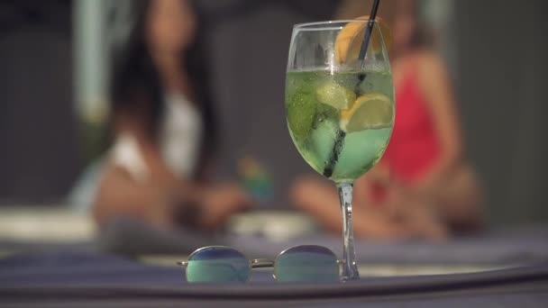 Cocktail fresco e óculos de sol no fundo de duas garotas de maiô. Foco no primeiro plano . — Vídeo de Stock