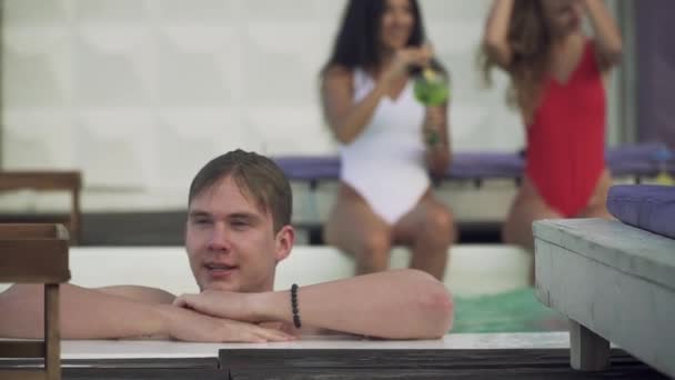 Jovem molhado relaxando na piscina ao ar livre no fundo de duas meninas bonitas. Uma companhia de jovens passa o tempo de lazer na piscina . — Vídeo de Stock