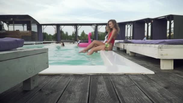 Красиві дівчата розважаються у відкритому басейні, створюючи величезні бризки води тонкими стрункими ногами — стокове відео