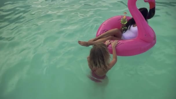 Draufsicht von niedlichen lockigen Blondine Spaß beim Rollen aufblasbaren Flamingo mit Coctail trinken heiße Brünette auf dem azurblauen Wasser. Freizeit und Spaß Mädchen. — Stockvideo