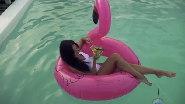 Two girlfriends in bathing suits have fun in the pool and drink cocktails. One girl with dark hair sits on an inflatable pink flamingo, the second with curly hair stands nearby. Leisure and fun girls. — Stock Video