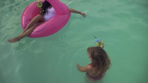 Две красивые девушки в купальниках веселятся в бассейне и пьют коктейли. Одна девушка с темными волосами сидит на надувном розовом фламинго, вторая с вьющимися волосами стоит рядом . — стоковое видео