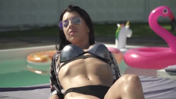 Jolie femme caucasienne avec un corps parfait en maillot de bain argenté est bains de soleil posé près de la piscine. Loisirs de mignonne dame en bikini et lunettes de soleil . — Video