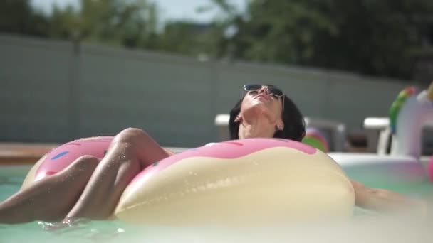サングラスのきれいな女性は、ピンクのゴム製のリングの上に横たわるプールで日光浴を楽しんでいます。ビキニでの孤独な女性のレジャー. — ストック動画
