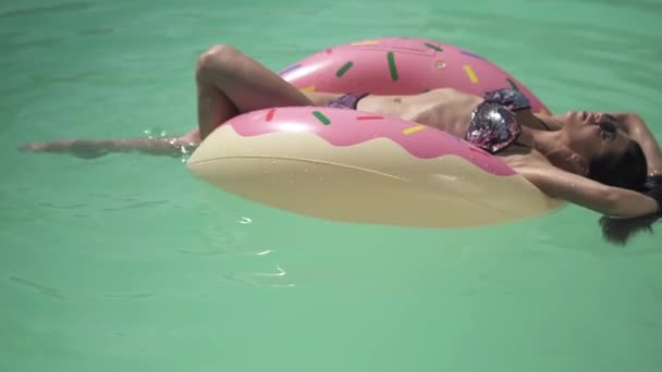 Güneş gözlüklü Şirin genç kadın üzerinde pembe Kauçuk halka yalan havuzu yüzme güneş sahiptir. Bir bikini yalnız kadın eğlence. — Stok video