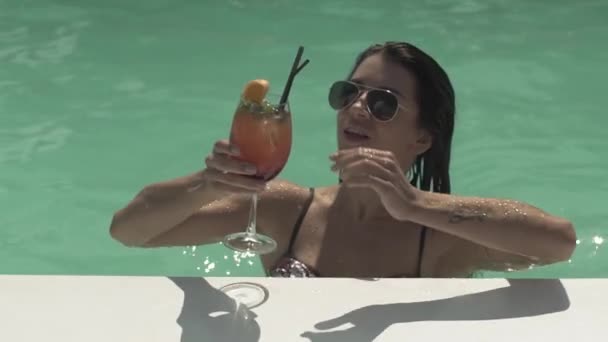 Брюнетка в солнечных очках пьет свой коктейль, стоя в бассейне. Отдых одинокой дамы в бикини . — стоковое видео