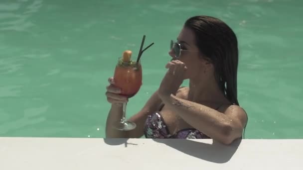 Carina giovane donna in occhiali da sole sta bevendo il suo cocktail in piedi in piscina. Tempo libero di signora solitaria in bikini . — Video Stock