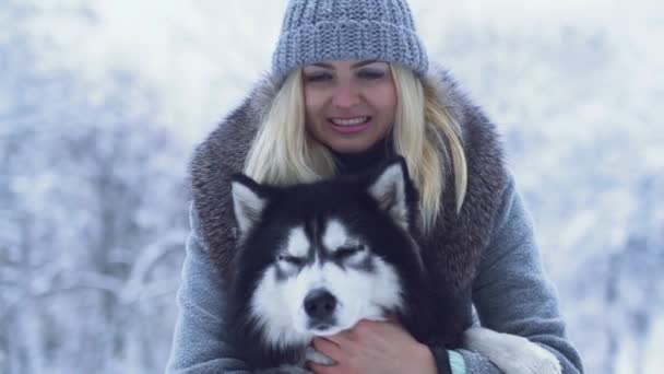 Porträt einer hübschen jungen Frau, die im winterlichen Schneehintergrund einen flauschigen sibirischen Husky hält und umarmt. Hund beim Winterspaziergang im Wald. — Stockvideo