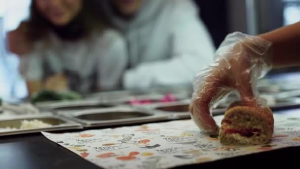 Junge glückliche Menschen, die verliebt zusehen, wie die männlichen Hände in Einweghandschuhe eingewickelt Sandwich in Packpapier. Junges Paar im Fast-Food-Laden. — Stockvideo