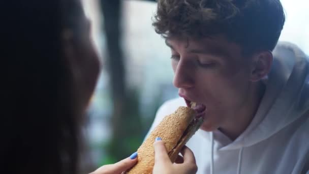 Meisje vrolijk en speels voedt haar lachende vriendje met boterham en dan zachtjes veegt zijn mond. Jong koppel in de fast-food winkel. — Stockvideo