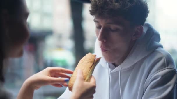 Ragazza allegramente e giocosamente nutre il suo fidanzato sorridente con panino. Coppia giovane nel fast food. Coppia giovane nel fast food . — Video Stock