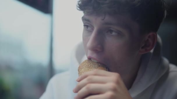 Портрет молодого привлекательного парня, который ест сэндвич и думает в кафе — стоковое видео