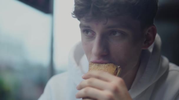 Retrato de jovem atraente smilling cara comer sanduíche e pensar na loja de fast food — Vídeo de Stock