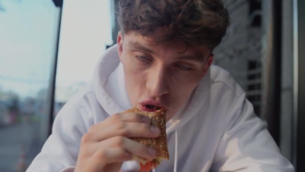 Портрет молодого привабливого усміхненого хлопця, який їсть бутерброд і думає в магазині фаст-фуду — стокове відео