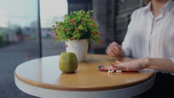 Девушка отбрасывает витаминные таблетки рукой со стола и берет зеленое вкусное яблоко, сидя на террасе открытого кафе — стоковое видео