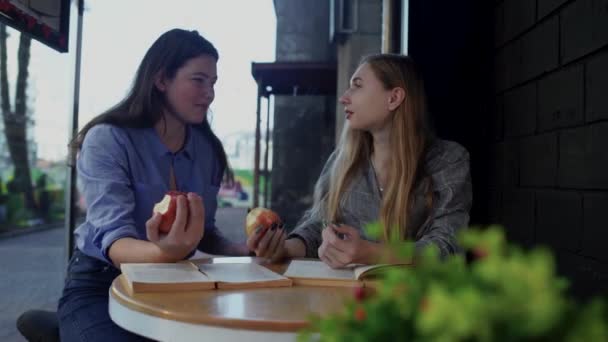 Dos chicas sentadas en una mesa en un café de la calle comiendo manzanas y discutiendo un libro. Las niñas llevan un estilo de vida saludable y disfrutan hablando entre ellas. novias morder una manzana y mostrar los pulgares hacia arriba . — Vídeos de Stock