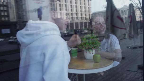 Skott av en ung uy och flickan för ett datum vid ett café bord genom ett glasfönster från gatan. Ungt par i kärlek vilar på ett café tillsammans. — Stockvideo