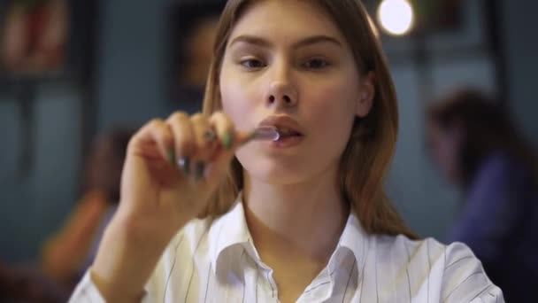 Retrato de una joven con una camisa blanca sentada en un café sosteniendo una pluma cerca de los labios . — Vídeos de Stock