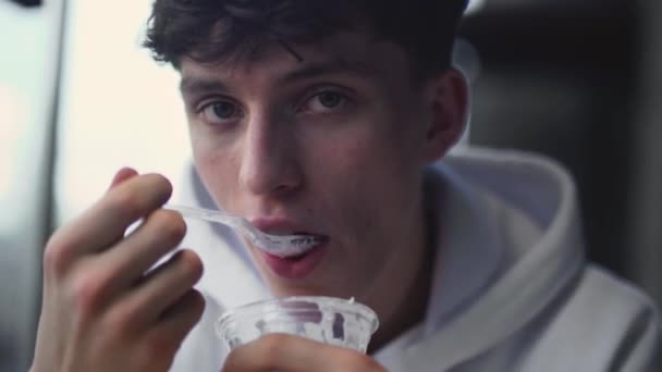 Портрет молодого парня, который ест вкусный сладкий йогурт и предлагает с красивой улыбкой попробовать его тоже — стоковое видео